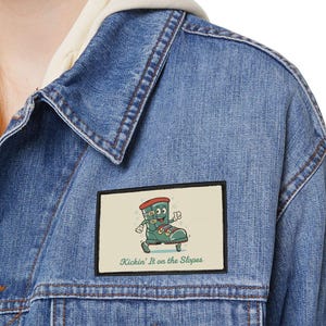 Op de afbeelding: Een lichtblauwe spijkerjas met een patch met een cartoonlaars op een skateboard. De patch heeft de tekst "Kickin' It on the Slopes". De jas heeft een knoopsluiting en twee borstzakken.