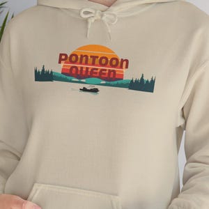 Pode incluir: Um moletom com capuz bege com um bolso frontal. O moletom apresenta um gráfico com as palavras "Pontoon Queen" em vermelho, um pôr do sol, árvores e um barco na água. O moletom é feito de um material macio.