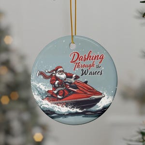 Könnte beinhalten: Runder Weihnachtsschmuck mit Santa Claus auf einem roten Jet-Ski, der durch Wellen fährt. Der Schmuck trägt den Text "Dashing Through the Waves" in roter Schrift. Der Hintergrund ist hellblau.