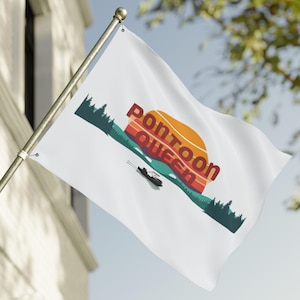 Pode incluir: Bandeira branca com as palavras "Pontoon Queen" em vermelho e laranja, com um desenho de pôr do sol e um barco pontão num lago. A bandeira está a ondular ao vento, montada num mastro prateado.