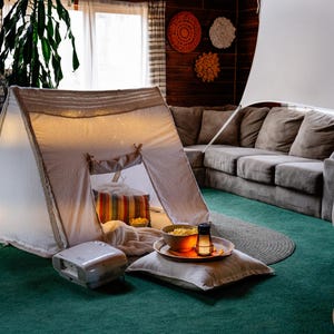 May include: A cozy indoor setup featuring a white teepee tent with string lights, a projector, and a bowl of popcorn. A large gray sectional sofa and a screen are set up for a home theater experience. The room has a green carpet and decorative wall hangings.