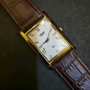 Peut inclure: Montre Seiko Quartz dorée de forme rectangulaire avec un cadran blanc et des chiffres romains. Les aiguilles sont noires et le bracelet est en cuir marron avec un motif crocodile. Design classique.