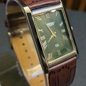 Pode incluir: Um relógio Seiko Quartz retangular com mostrador verde e ponteiros e marcadores dourados. O relógio tem uma pulseira de couro castanho. O mostrador exibe algarismos romanos e o logotipo Seiko.