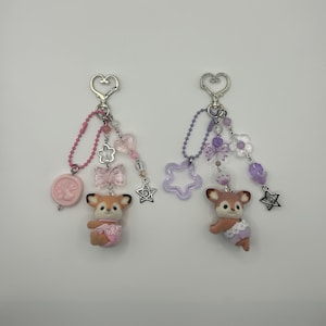 May include: Two keychains featuring small deer figurines. The left keychain has pink beads, a pink circular charm, a clear bow, and a heart-shaped clasp. The right keychain has purple beads, a purple star-shaped charm, a clear flower, and a heart-shaped clasp.