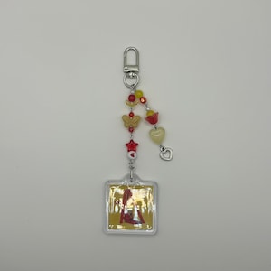May include: A clear acrylic keychain with a square pendant featuring a colorful image. The keychain has a silver clasp and is adorned with red, yellow, and clear beads, including butterfly and heart shapes.