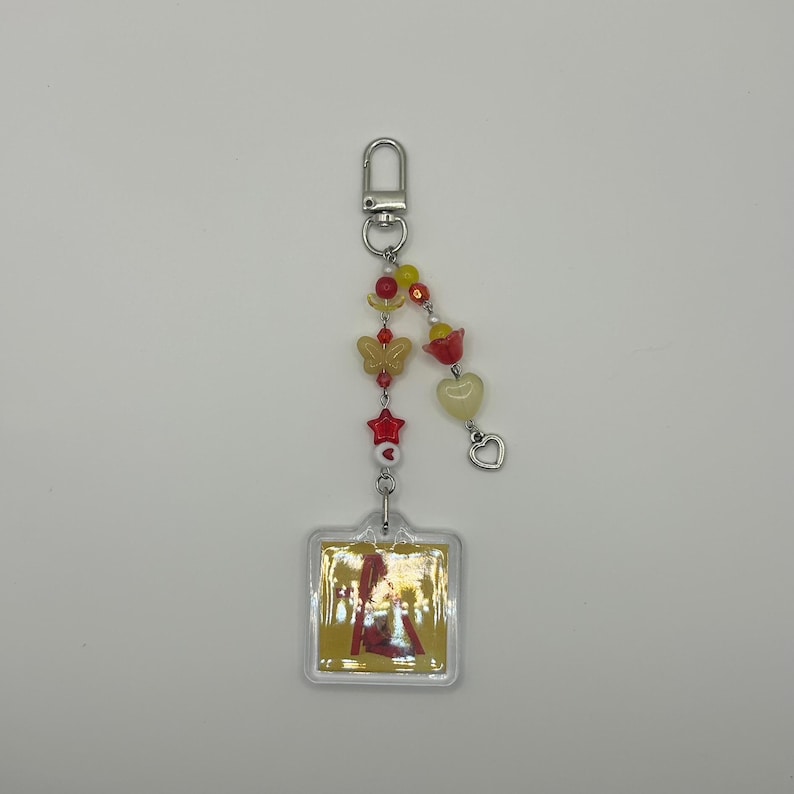 May include: A clear acrylic keychain with a square pendant featuring a colorful image. The keychain has a silver clasp and is adorned with red, yellow, and clear beads, including butterfly and heart shapes.