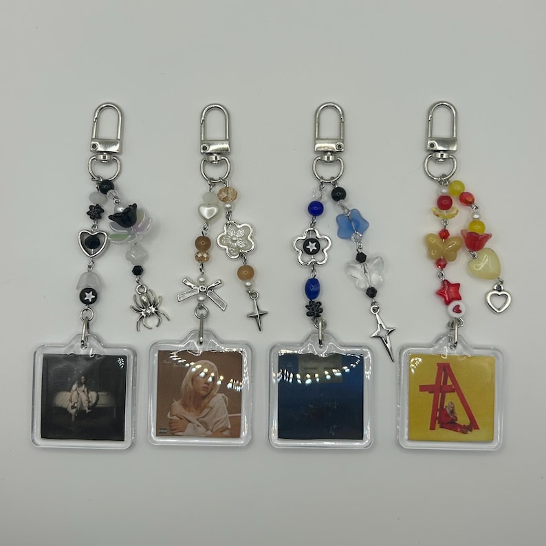 May include: Four keychains with silver clasps, each featuring a small, square image. The keychains have decorative beads, charms, and pendants in various colors. The images appear to be album art or similar graphics.