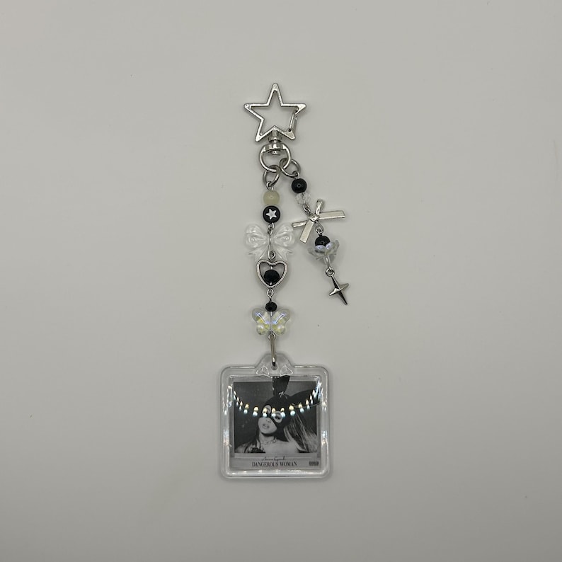 May include: A silver keychain with a star-shaped clasp, adorned with black and white beads, bows, and charms. A small, clear square pendant features a black and white image with the text "DANGEROUS WOMAN".
