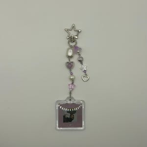 May include: A silver keychain with a star-shaped clasp, adorned with a string of beads in shades of purple, white, and silver. The keychain features a small, clear square with a photo of a deer. The design includes heart and star charms.