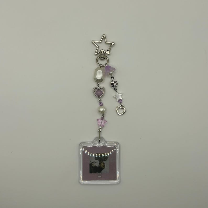 May include: A silver keychain with a star-shaped clasp, adorned with a string of beads in shades of purple, white, and silver. The keychain features a small, clear square with a photo of a deer. The design includes heart and star charms.