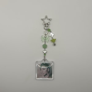 May include: A clear acrylic keychain with a photo of a person. The keychain has a silver star-shaped clasp and is adorned with green beads, flowers, and a bow. The photo is square and has a clear border.