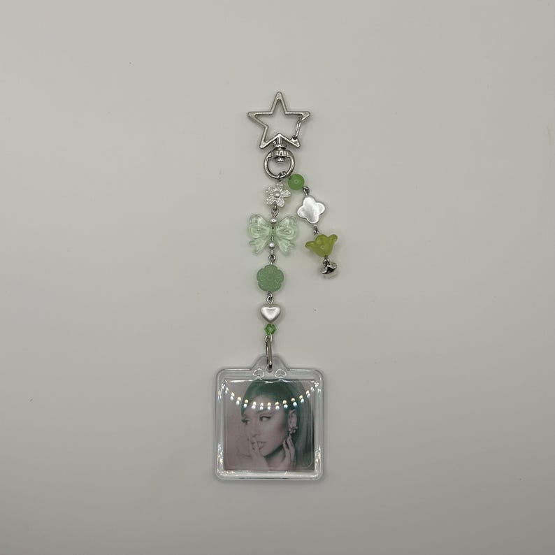 May include: A clear acrylic keychain with a photo of a person. The keychain has a silver star-shaped clasp and is adorned with green beads, flowers, and a bow. The photo is square and has a clear border.