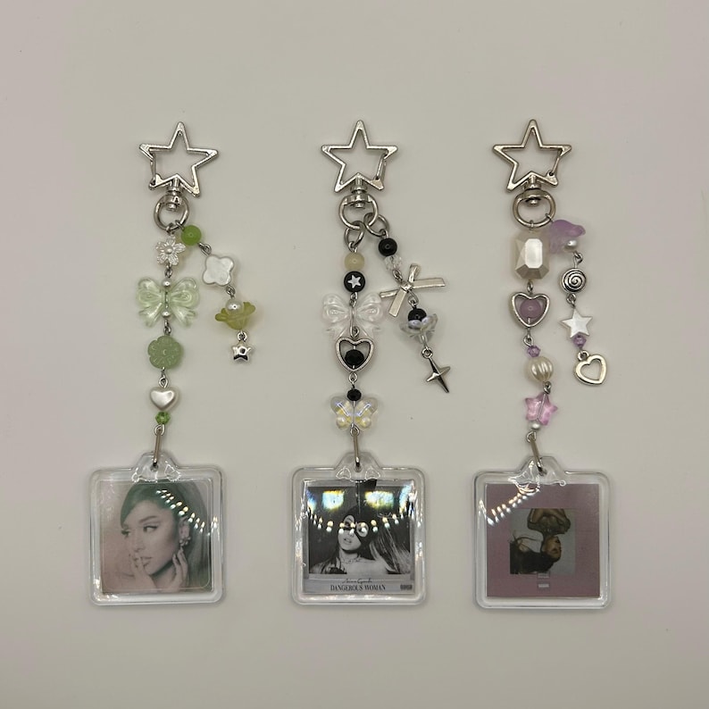 May include: Three decorative keychains with star-shaped clasps. Each keychain features a clear square pendant with a photo, and a beaded charm with various shapes and colors, including butterflies, hearts, and stars. The keychains are silver in color.