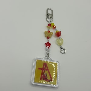 May include: A clear acrylic keychain with a yellow and red design. The keychain features a decorative beaded chain with butterfly, star, and heart charms. The main charm has a yellow background with a red design and the letter "A".