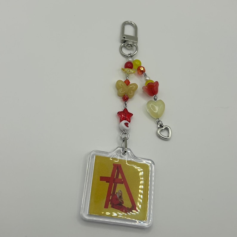 May include: A clear acrylic keychain with a yellow and red design. The keychain features a decorative beaded chain with butterfly, star, and heart charms. The main charm has a yellow background with a red design and the letter "A".