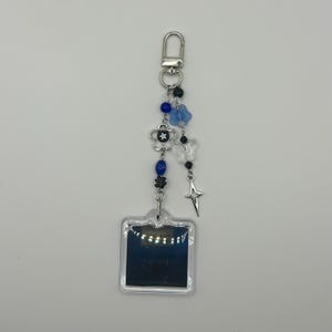 May include: A silver keychain with a clear, square charm. The keychain features blue and black beads, a silver flower, butterfly charms, and a star charm. The keychain is attached to a silver clasp.