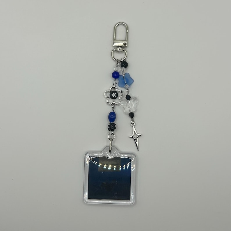 May include: A silver keychain with a clear, square charm. The keychain features blue and black beads, a silver flower, butterfly charms, and a star charm. The keychain is attached to a silver clasp.