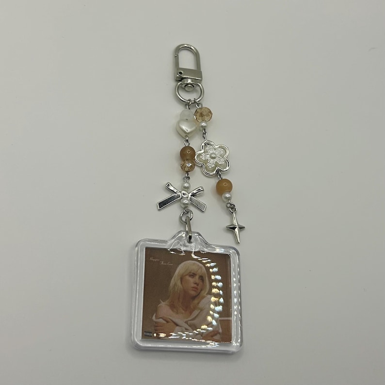 May include: A clear acrylic keychain featuring a photo of a person with blonde hair. The keychain has a silver-colored clasp and is adorned with beads, a flower charm, a bow charm, and a star charm. The beads are in shades of white, brown, and silver.