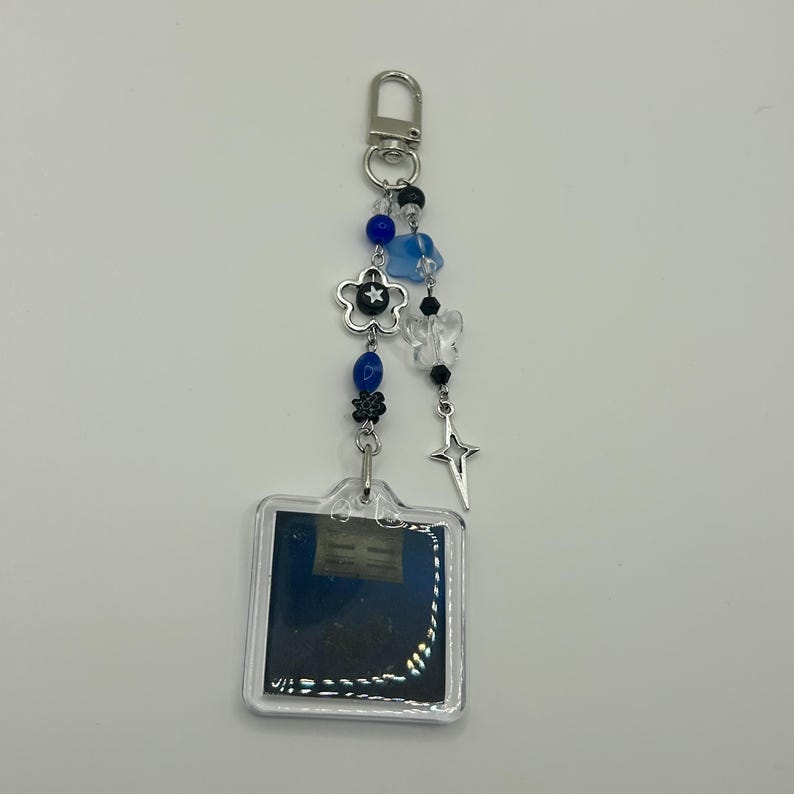 May include: A clear acrylic keychain with a silver clasp and decorative charms. The charms include blue and clear beads, a silver flower, a star, and a small square with a dark blue background.