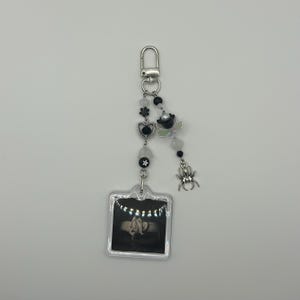 May include: A silver keychain with a clear, square charm featuring a black and white photo. The keychain has a beaded strand with black, white, and iridescent beads, a heart, a star, and a spider charm.