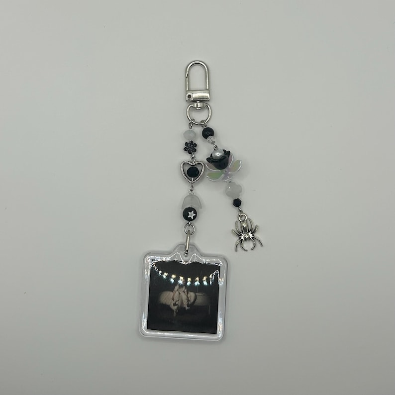May include: A silver keychain with a clear, square charm featuring a black and white photo. The keychain has a beaded strand with black, white, and iridescent beads, a heart, a star, and a spider charm.