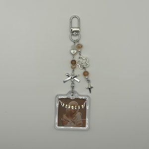 May include: A decorative keychain with a silver clasp, adorned with beads, a flower charm, a bow, and a star. A clear, square pendant displays a photo of a person. The keychain is a mix of silver, beige, and clear elements.