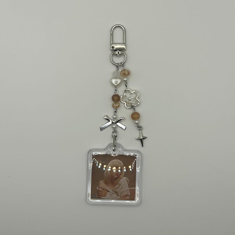 May include: A decorative keychain with a silver clasp, adorned with beads, a flower charm, a bow, and a star. A clear, square pendant displays a photo of a person. The keychain is a mix of silver, beige, and clear elements.