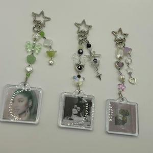 May include: Three keychains with square photo holders. Each keychain has a star-shaped clasp and is adorned with beads, charms, and a small photo. The keychains feature different color schemes, including green, black, and pink, with various charms.