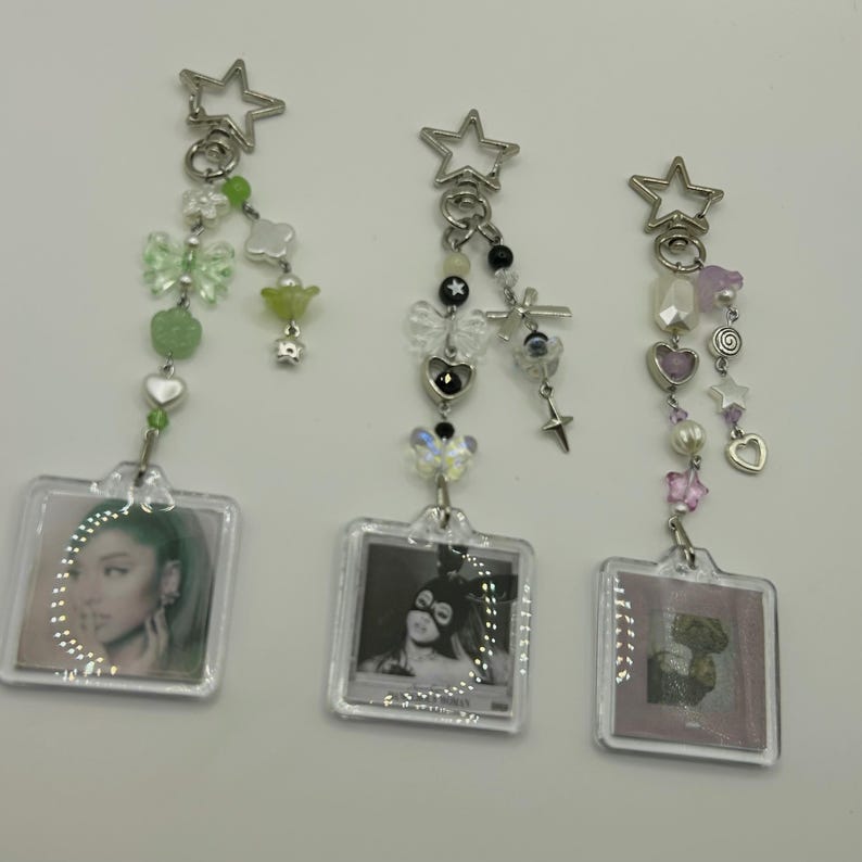 May include: Three keychains with square photo holders. Each keychain has a star-shaped clasp and is adorned with beads, charms, and a small photo. The keychains feature different color schemes, including green, black, and pink, with various charms.