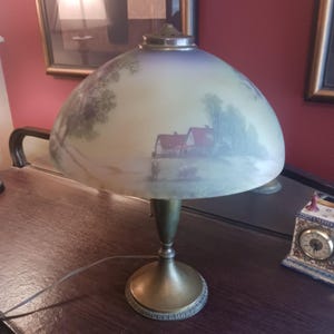 Può includere: Una lampada da tavolo vintage con un paralume in vetro dipinto raffigurante un paesaggio con case e alberi. La lampada ha una base in ottone e un paralume curvo a forma di cupola. La lampada è su una superficie di legno.