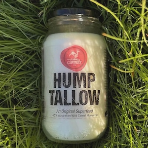 Puede incluir: Un tarro de vidrio lleno de una sustancia blanca cremosa, etiquetado como "HUMP TALLOW" con el texto "An Original Superfood" y "100% Australian Wild Camel Hump Fat." El tarro tiene una tapa negra y un logotipo rojo y blanco. El tarro está sobre hierba verde.
