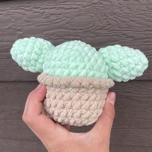 Pu&ograve; includere: Un cactus all'uncinetto fatto a mano in un vaso beige con cuscinetti di cactus verde chiaro. Il cactus &egrave; tenuto in mano su uno sfondo di legno marrone. L'uncinetto ha un aspetto strutturato e morbido.