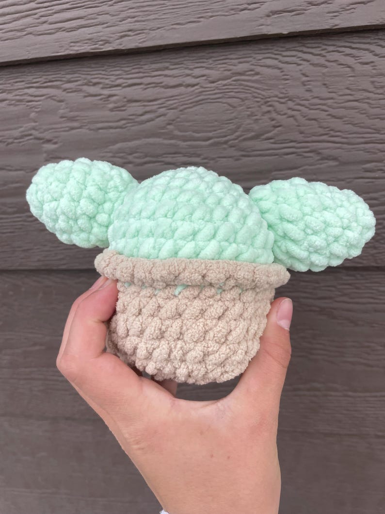 Pu&ograve; includere: Un cactus all'uncinetto fatto a mano in un vaso beige con cuscinetti di cactus verde chiaro. Il cactus &egrave; tenuto in mano su uno sfondo di legno marrone. L'uncinetto ha un aspetto strutturato e morbido.