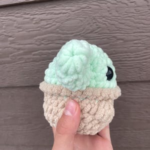Pu&ograve; includere: Una figura all'uncinetto fatta a mano a forma di personaggio con grandi orecchie e testa rotonda. La figura ha una testa verde chiaro e una base beige. Gli occhi sono neri. La figura &egrave; tenuta in mano.