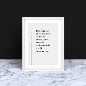 Op de afbeelding: Een ingelijste print met een witte rand toont de tekst "De grootste guru mantra is: deel nooit je geheimen met iemand. Het zal je vernietigen." De achtergrond is een zwarte muur en een oppervlak met marmerpatroon.