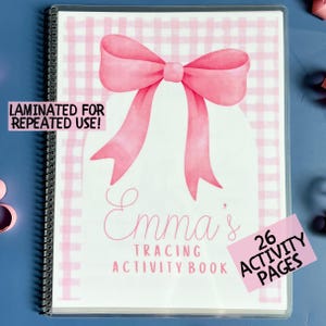May include: A spiral-bound activity book with a pink bow illustration on the cover. The cover has the text "Emma's Tracing Activity Book" and "Laminated for Repeated Use!" A pink sticker reads "26 Activity Pages".