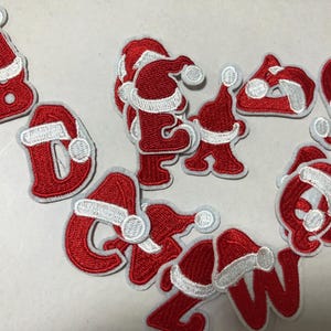 May include: A collection of red and white embroidered patches in the shape of letters and Santa hats. The letters appear to spell out "Christmas" in a festive, holiday-themed design. The patches are on a light gray background.