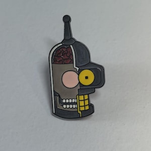 Pode incluir: Um pin com um design estilizado da cabeça de um robô, dividida em duas. Um lado mostra o cérebro, o outro o rosto com um olho amarelo e uma bochecha rosa. O pin é em tons de cinza, preto e bege, com um acento amarelo.