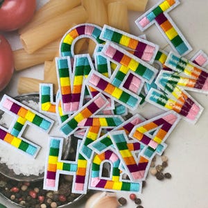 May include: Embroidered rainbow-colored alphabet letters with white borders. The letters are scattered on a surface with tomatoes, pasta, and spices. The letters are in various colors, including pink, yellow, green, and blue.