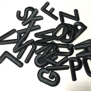 Op de afbeelding: Een verzameling zwarte, geborduurde stoffen letters verspreid op een witte ondergrond. De letters zijn in een schreefloze lettertype en lijken individuele patches of applicaties te zijn, geschikt voor knutsel- of personalisatieprojecten.