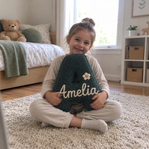 May include: A dark green, textured, letter-shaped cushion in the form of the letter A. The cushion features a small, white, five-petaled flower accent and the name Amelia embroidered in white cursive script across the front surface.