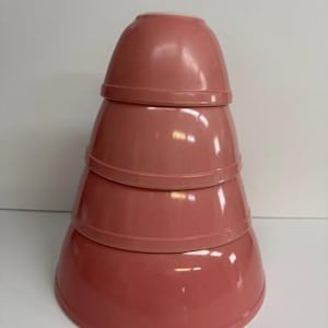 May include: Four rose-coloured nesting bowls stacked together, creating a conical shape. The bowls are arranged from largest at the base to smallest at the top. They have a glossy finish and are designed for storage.
