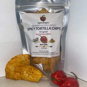 May include: A clear bag of TopNotchPeppers Spicy Tortilla Chips, Original Red Habanero Edition, with two chips in front. The bag has a label with text and illustrations of garlic, a red pepper, and spices. Two red habanero peppers are next to the bag.