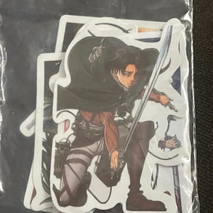 Puede incluir: Un conjunto de pegatinas de estilo anime que representan a un personaje con una espada, vestido con una chaqueta marrón, pantalones blancos y una capa negra. Las pegatinas están en una bolsa de plástico transparente.