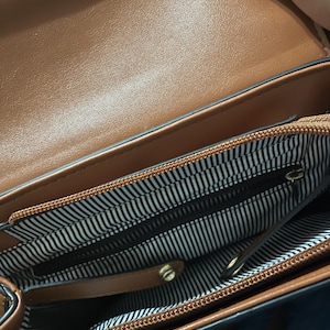 May include: Open view of a brown handbag with a top handle. The interior features a black and white striped lining and multiple compartments. A gold-colored clasp and zipper are visible.