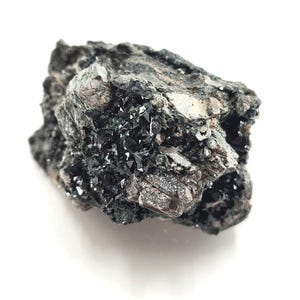 May include: A dark gray mineral specimen with clusters of small, shiny, black crystals. The rough, textured surface of the rock has areas of lighter gray and silver. The mineral sits on a white background.