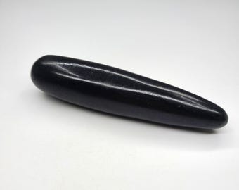 May include: A smooth, solid black stone wand with a polished finish. The object has an elongated, cylindrical shape that tapers to a rounded point at one end and features a blunt, rounded base at the other, presenting a uniform dark surface.