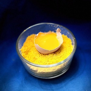 May include: A clear glass bowl filled with yellow crumbs, with an eggshell containing a bright yellow yolk resting on top. The background is a deep blue.