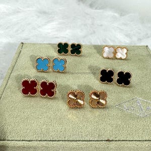 May include: A selection of clover-shaped stud earrings in various colours, including red, turquoise, green, black, white, and gold. The earrings are displayed on a green surface.
