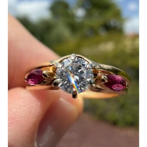 May include: A gold ring featuring a large, clear, round-cut diamond at its centre. Two smaller, oval-shaped, deep red gemstones are set either side of the main diamond. The ring's band is gold, with a gold and silver setting.